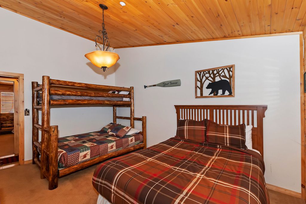 A cozy bedroom with a wooden bunk bed and a full-size bed with plaid comforters, a wood-paneled ceiling, and a bear-themed art piece on the wall in a cabin in Big Bear.