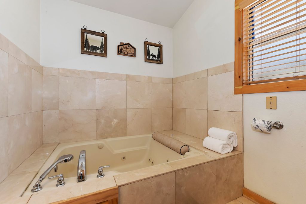 A large, tiled jetted bathtub with a small window, two rolled white towels on the side, and three small framed pictures on the wall in a bathroom in a Big Bear cabin.