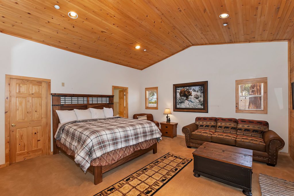 A large bedroom with a king-size bed, a small couch with a bear-themed pattern, a wooden trunk coffee table, and a wood-paneled ceiling in a Big Bear cabin.