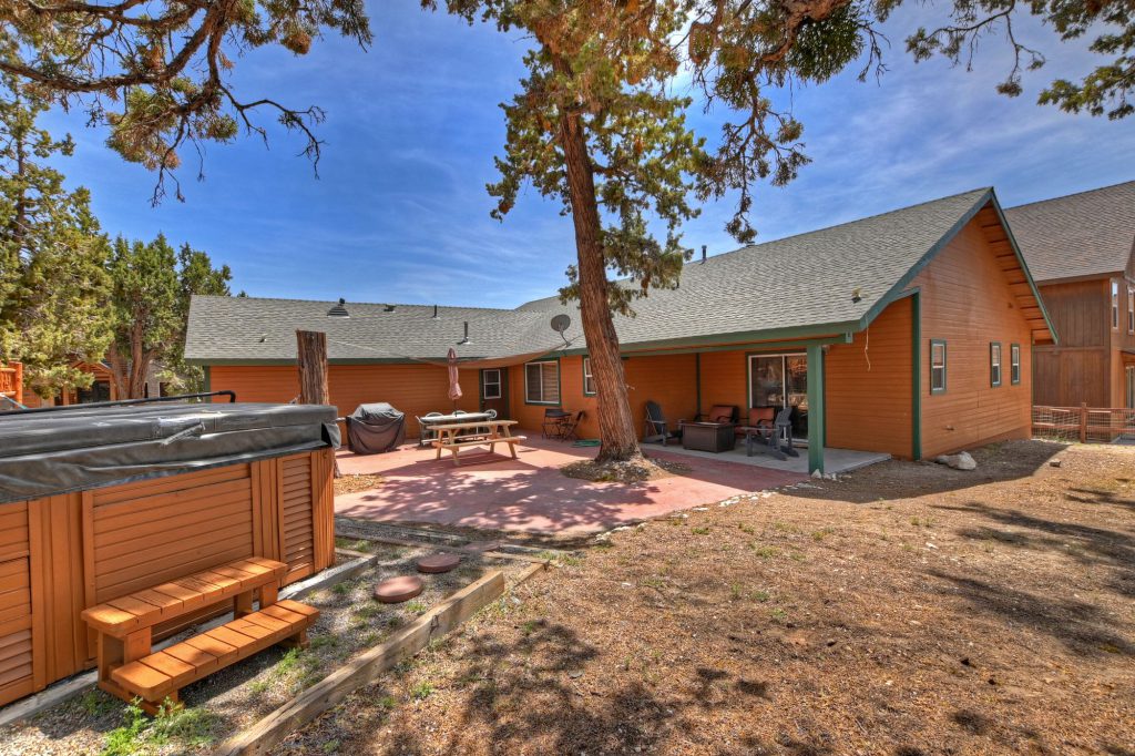 The backyard of a single-story wooden house with a hot tub on the left, a brick patio with a dining table, and a covered porch with a seating area, surrounded by a dirt and grass yard with tall pine trees in Big Bear.