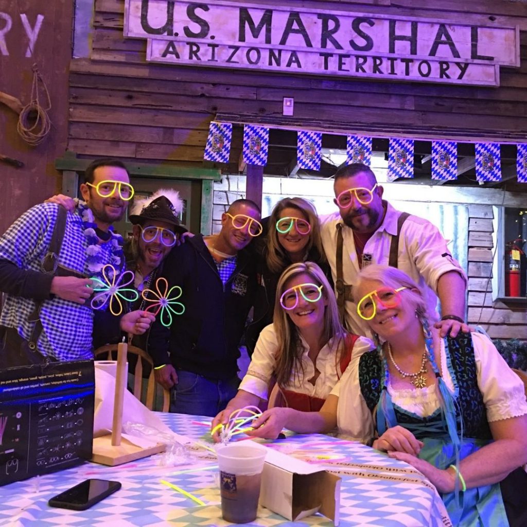 A group of people at the Big Bear Oktoberfest are wearing neon-rimmed glasses and posing for a picture, with some holding glowing toys.