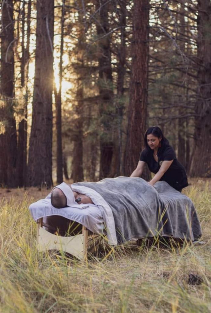A person receiving a massage outdoors on a portable massage table in a forest setting, covered with a gray blanket.
