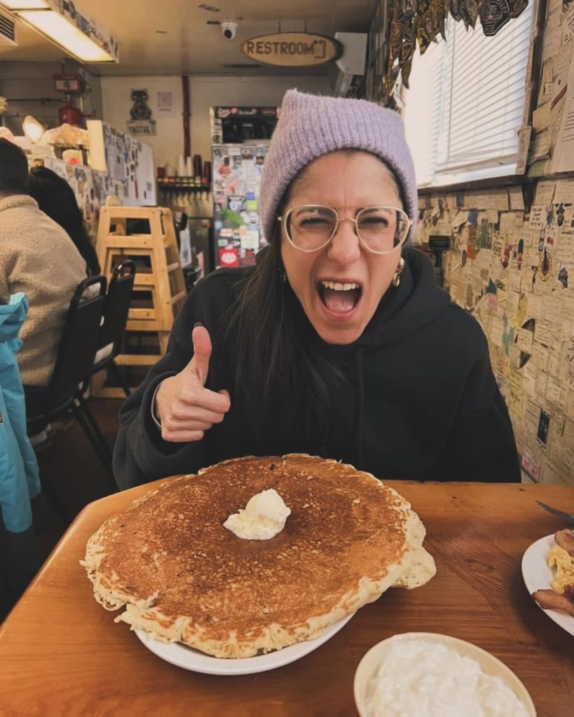 Person reacting excitedly while sitting in front of a giant pancake breakfast inside a cozy diner.