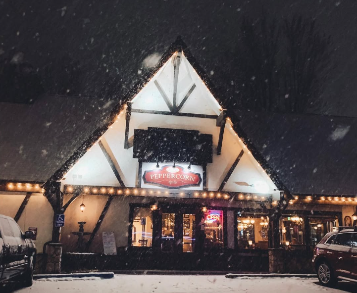 The exterior of a restaurant called Peppercorn Grille is shown at night in Big Bear, with snow falling and lights illuminating the building.