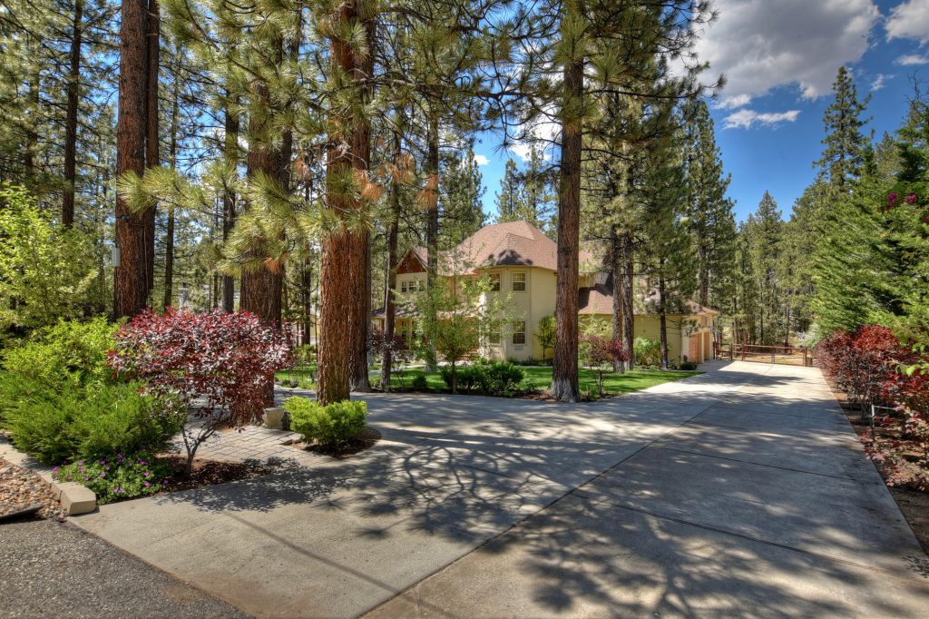 A large, multi-story cabin is nestled among tall pine trees in a wooded area at Big Bear, with a circular driveway and well-maintained landscaping in the foreground