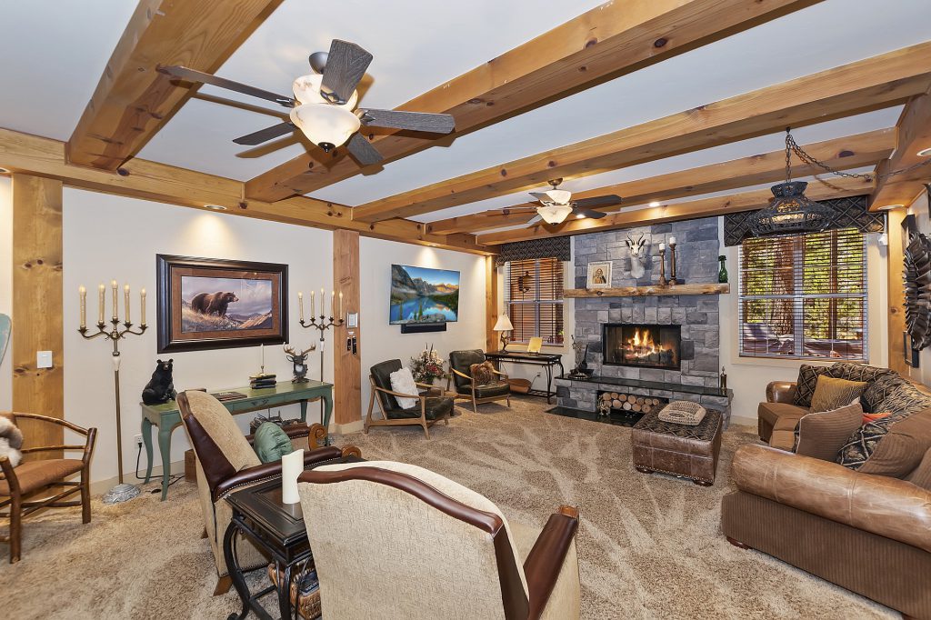 A cozy, rustic living room at Big Bear is shown, featuring a stone fireplace, exposed wood beams on the ceiling, comfortable seating, and multiple light fixtures.