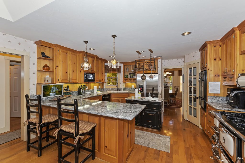 A rustic, spacious kitchen at Big Bear is shown, featuring granite countertops, light wood cabinetry, a breakfast bar with stools, and hardwood floors.