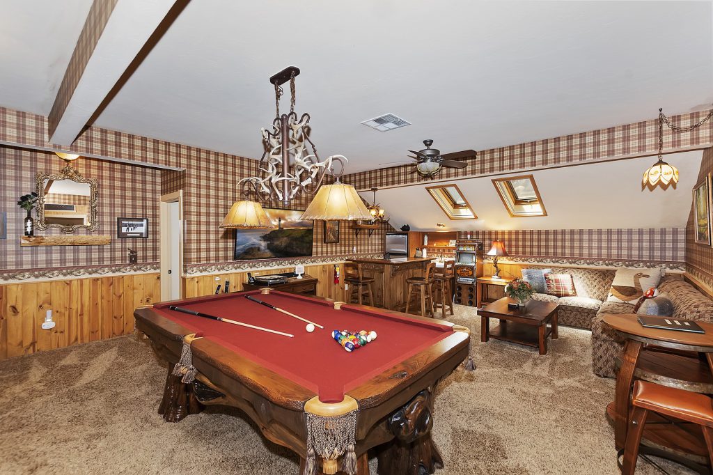 A rustic game room at Big Bear is shown, featuring a red-felt pool table, a wet bar, a sitting area, and a plaid wallpapered ceiling.