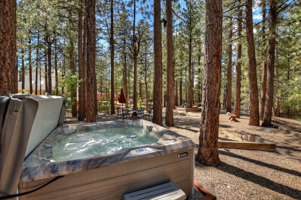 A bubbling hot tub is nestled in a clearing among tall pine trees at a Big Bear cabin, with a patio table and chairs visible in the background.