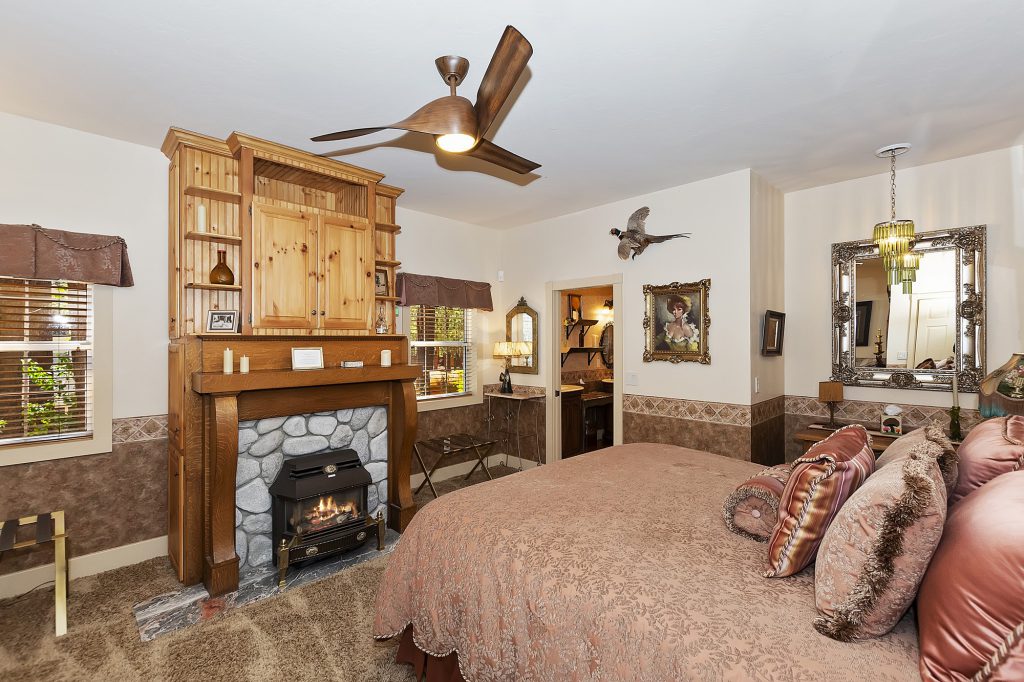 The interior of a master suite at Big Bear is shown, featuring a large bed, a stone fireplace with a wood mantle and built-in shelves, and a large ornate mirror.