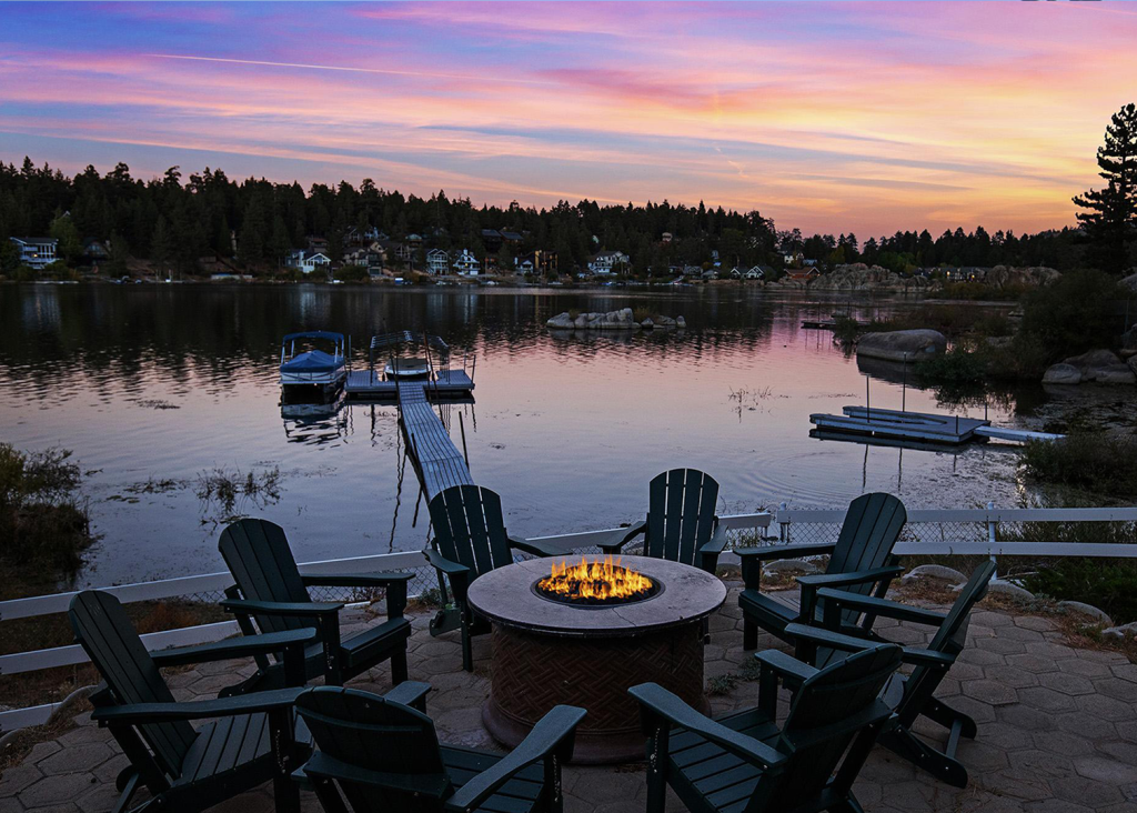 A fire pit with surrounding Adirondack chairs on a patio at Big Bear Lake, with a wooden dock and boats in the background at sunset.