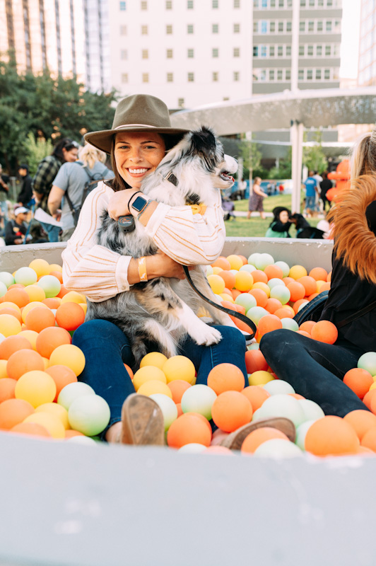 A smiling woman wearing a hat and her Australian Shepherd dog sit together in a ball pit filled with orange, yellow, and green balls.