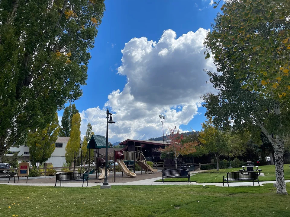Children's playground at Veterans Park in Big Bear surrounded by grassy areas, benches, and tall shade trees.