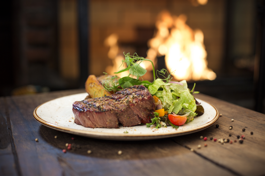 An image of a plate with a cooked steak, roasted potatoes, and a side salad, with a lit fireplace in the blurry background.