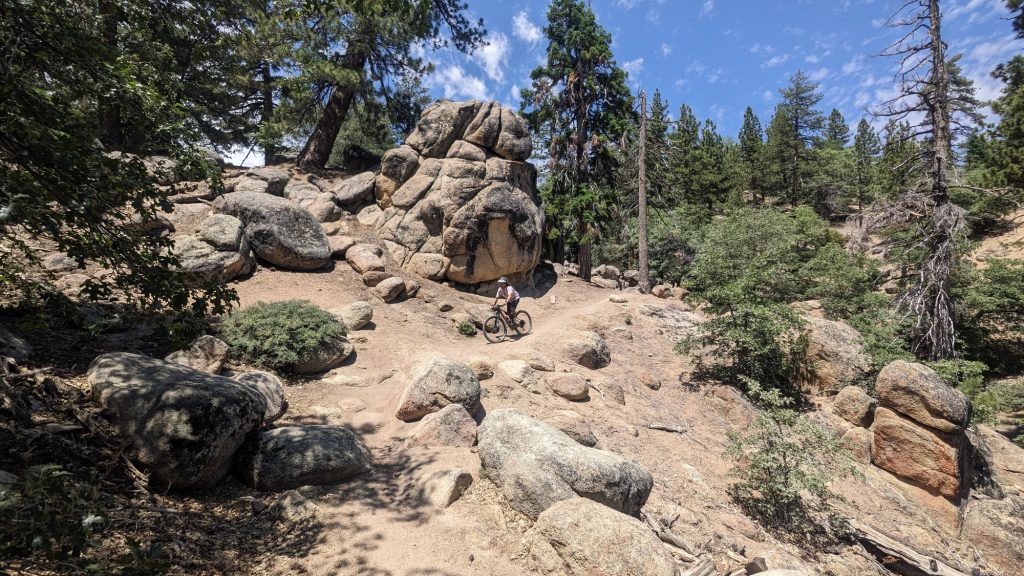A mountain biker riding through a rocky, technical trail with large boulders and pine trees, navigating rugged terrain in a forested mountain area.