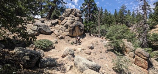 A mountain biker riding through a rocky, technical trail with large boulders and pine trees, navigating rugged terrain in a forested mountain area.