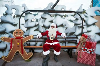 Santa Claus wearing sunglasses sitting on a ski chair lift with gingerbread man and presents around him