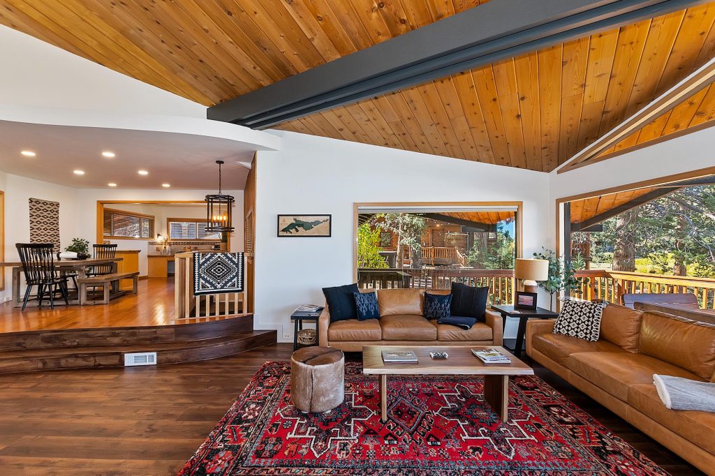Spacious mountain cabin living room with vaulted wood ceilings, leather sofas, open-concept layout, and large windows overlooking forest views.