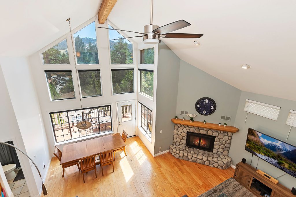 The interior of an A-frame cabin in Big Bear is shown from a high angle, with a dining table, a stone fireplace, and large windows that offer a view of the surrounding trees and mountains.
