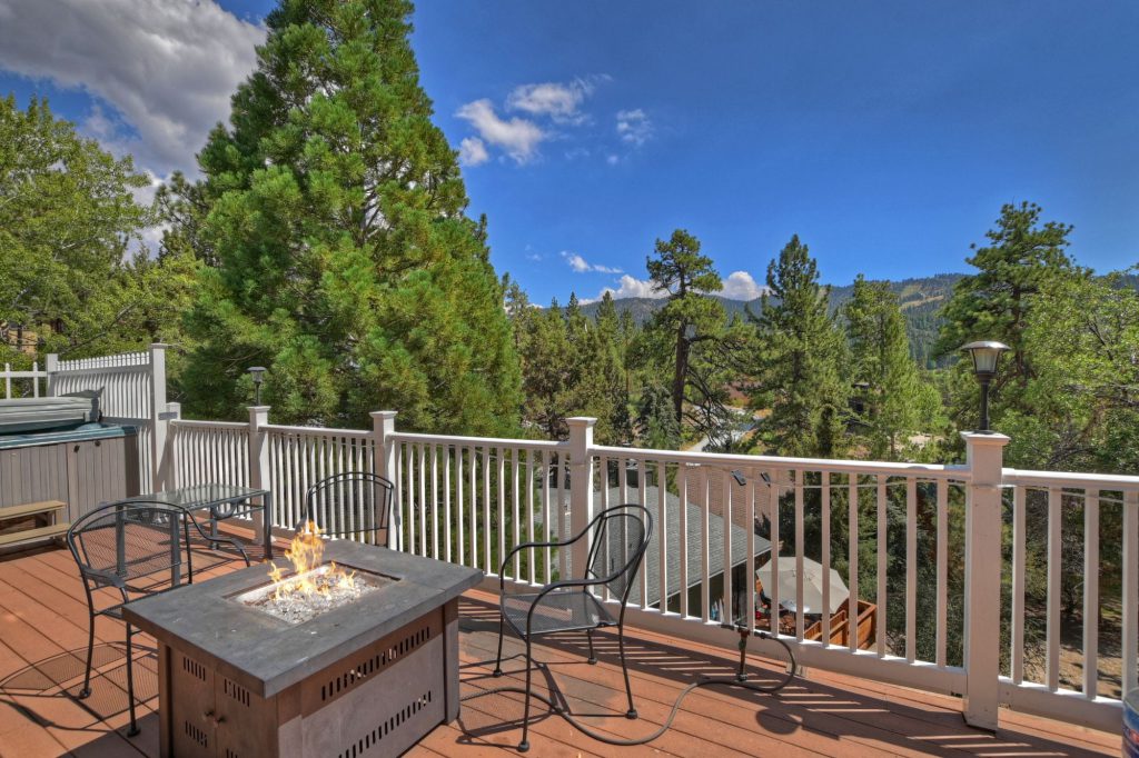 The deck of a Big Bear cabin is shown, featuring a hot tub, a metal fire pit with a flame, and several chairs, all surrounded by a white railing and pine trees.