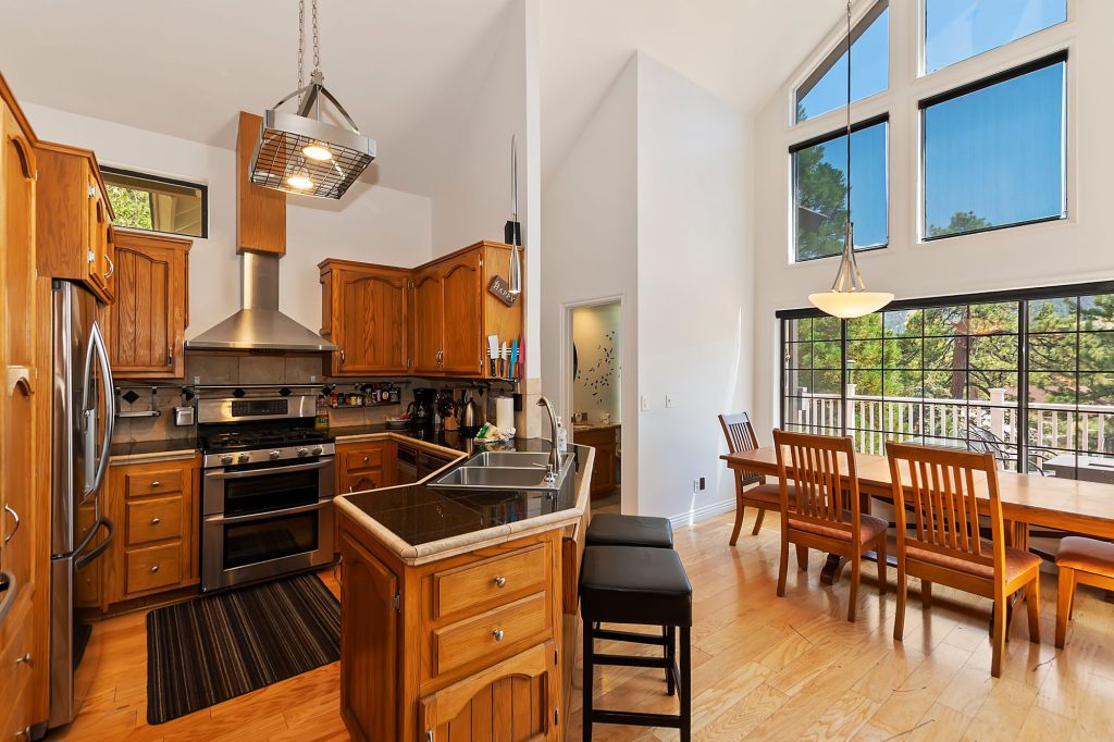 The kitchen and dining area of a Big Bear cabin is shown with wood cabinets, stainless steel appliances, a large dining table, and floor-to-ceiling windows overlooking a deck and trees.