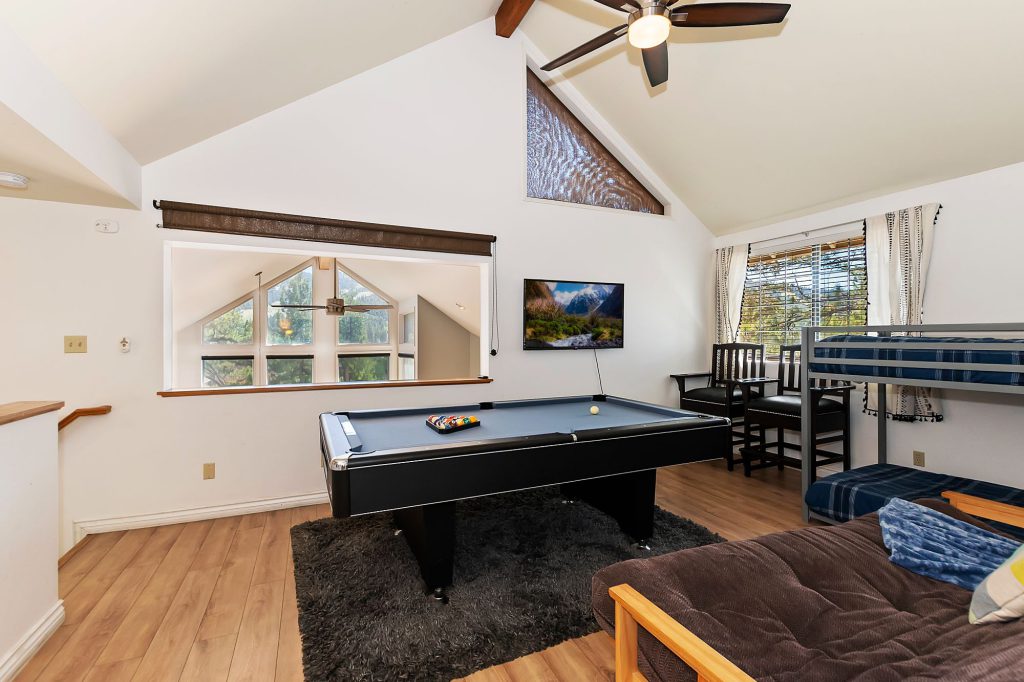 The loft of a Big Bear cabin is set up as a game room with a pool table on a grey rug, a futon, and a bunk bed, with a television mounted on the wall and a view into the living room below.