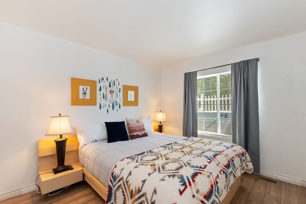 A bedroom in a Big Bear cabin is shown with a bed covered in a geometric-patterned blanket, two framed pictures on the wall, and a large window with grey curtains.