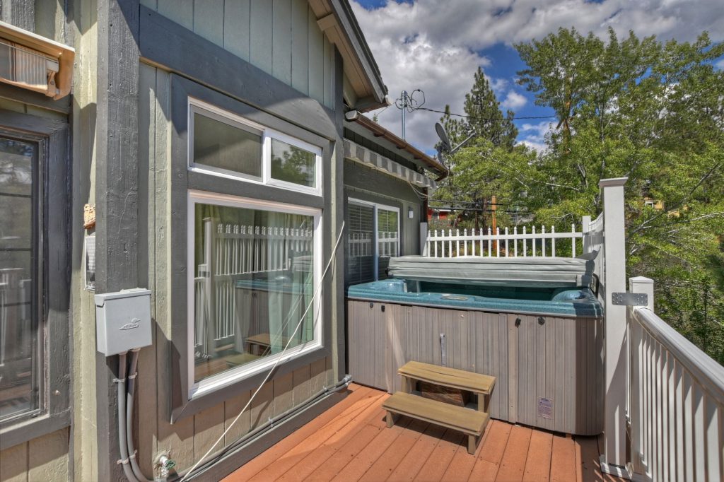 The exterior of a Big Bear cabin is shown, featuring a hot tub on a wooden deck with a white railing and a view of pine trees and a partly cloudy sky.
