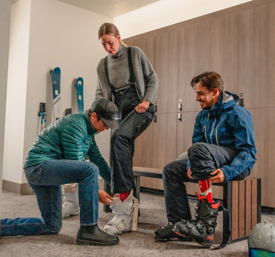 Two people trying on ski boots. One man sits in his boots on a bench while the other person is standing being assisted by a person kneeling helping them put on the boot.