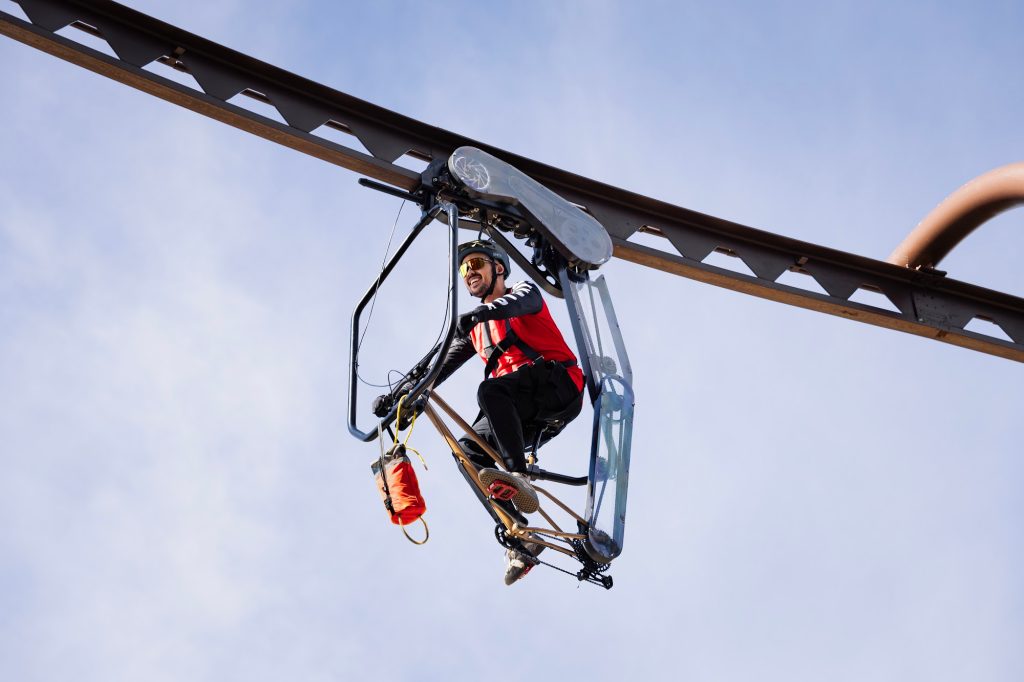 A person riding an elevated open-air aerial bike-style attraction suspended above the ground at SkyPark with clear sky background.