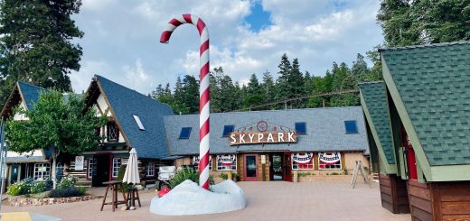 Wide view of SkyPark entrance plaza with alpine buildings, candy cane decoration, paved walkways, and visitors enjoying a mountain resort environment.