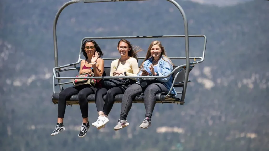 Three girls on a ski lift in Big Bear, California, surrounded by forested mountains, enjoying the adventure and views.