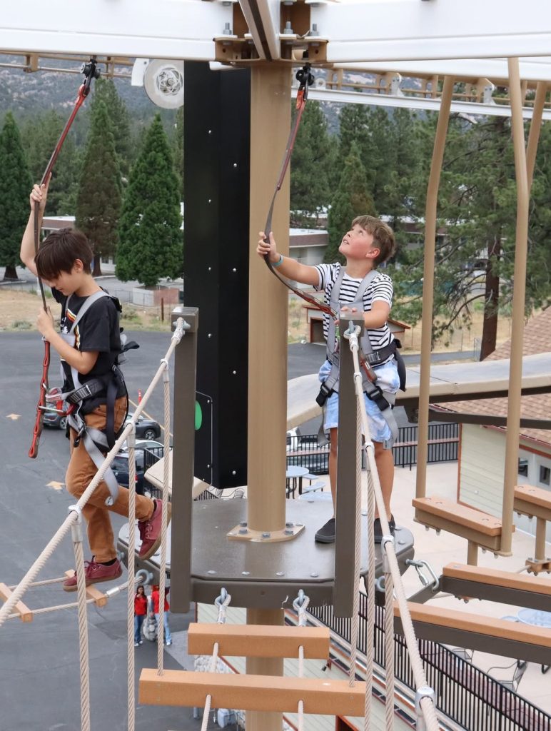 Children wearing safety harnesses navigating an aerial ropes course at an outdoor adventure park in Big Bear.