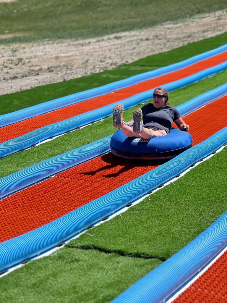 Person riding an inflatable tube down a summer tubing lane with artificial turf and colorful tracks in Big Bear.