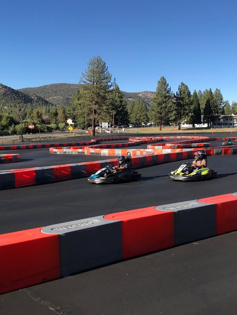 People racing go-karts on an outdoor track in Big Bear with mountain views and red-and-black safety barriers.