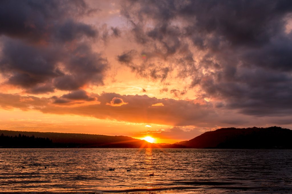 Dramatic orange sunset over Big Bear Lake with dark mountain silhouettes, colorful clouds, and reflections across the water