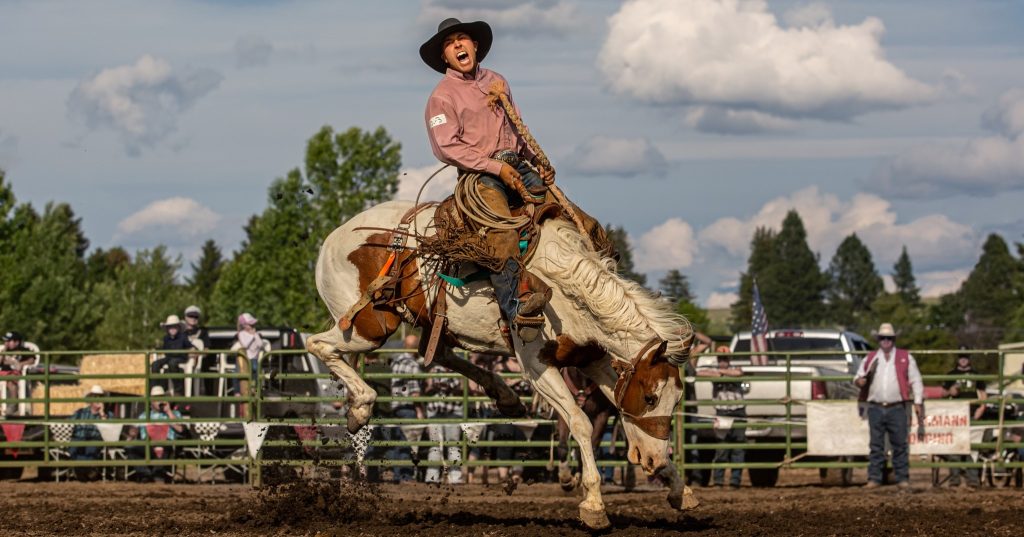 Cowboy riding a bucking horse during a rodeo event with spectators watching outdoors.