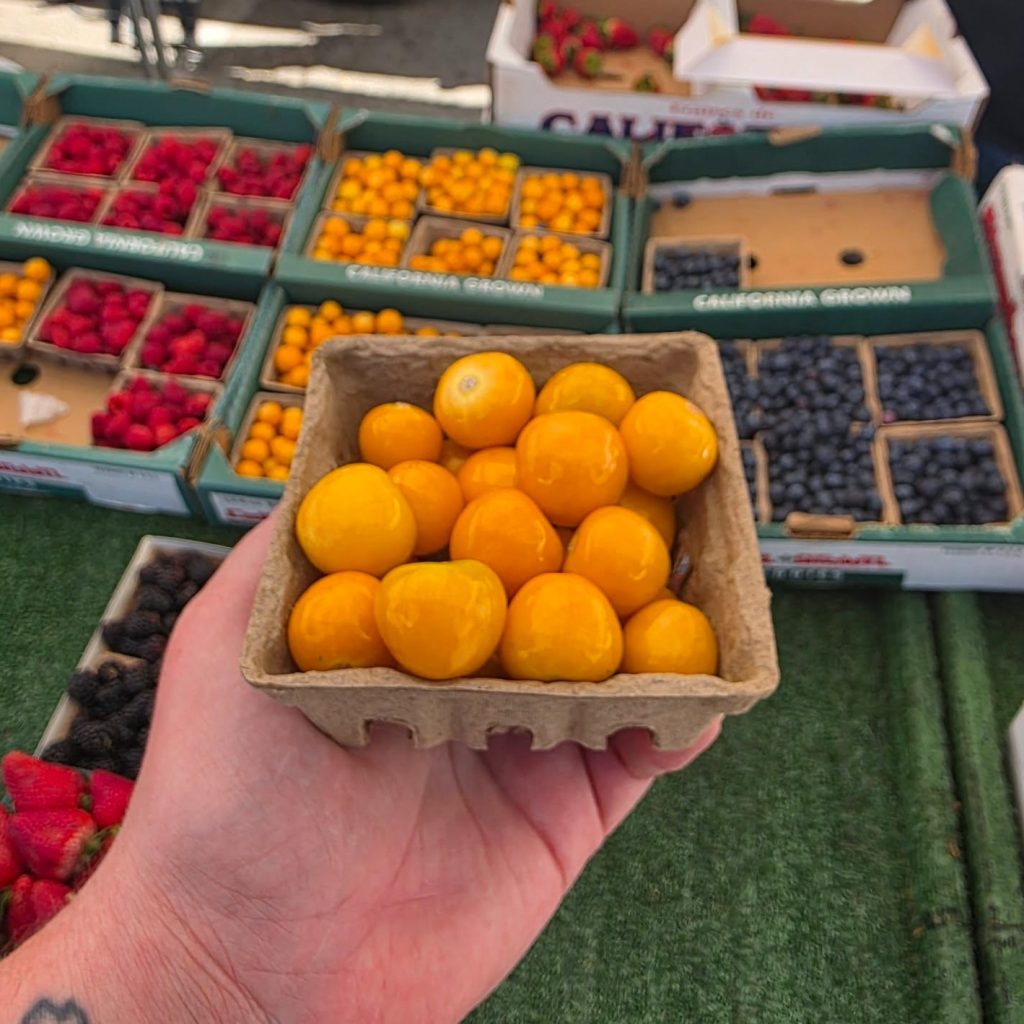 Fresh yellow citrus fruits displayed at a farmers market with colorful berry boxes in the background