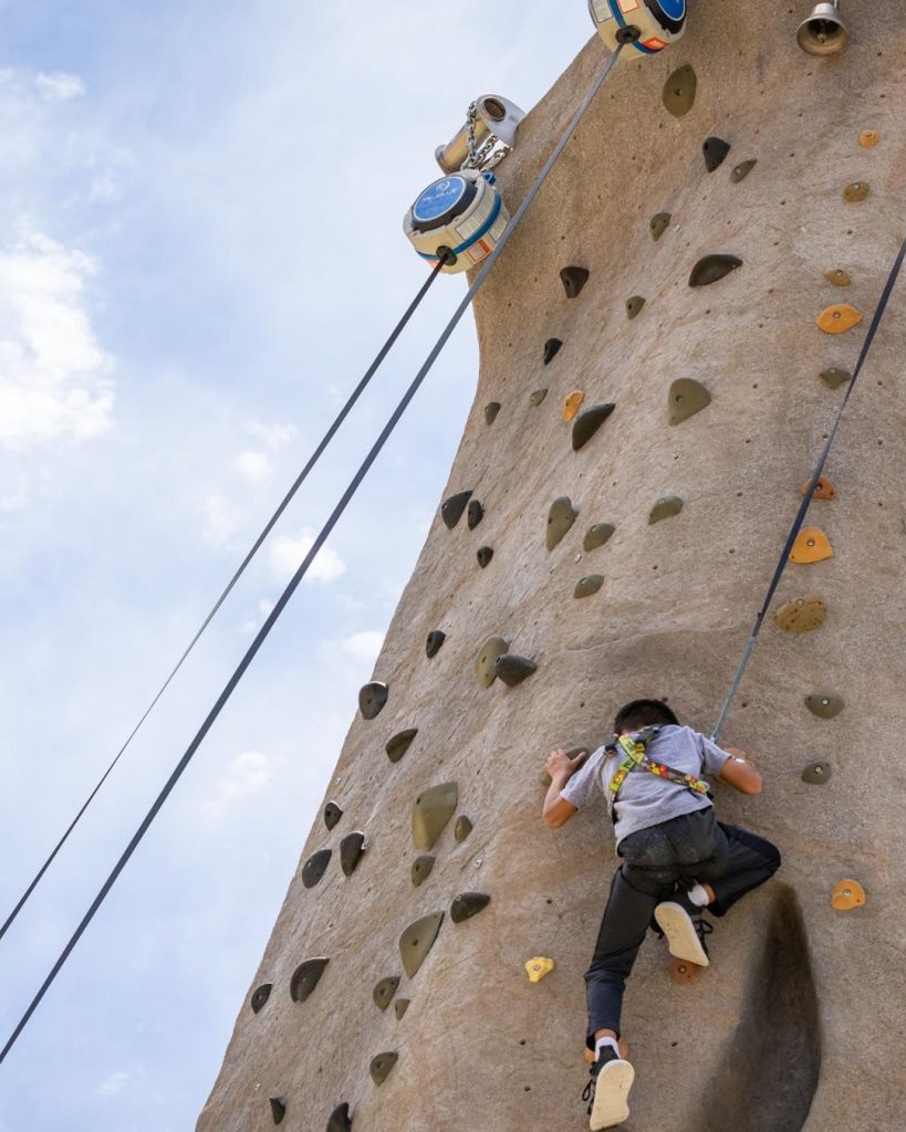 A child climbs the Snow Summit climbing wall using a safety harness on an outdoor adventure attraction in Big Bear.