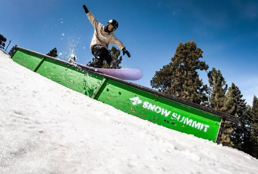 Snowboarder doing a trick on a green rail at Snow Summit in Big Bear, with snow and pine trees in the background.