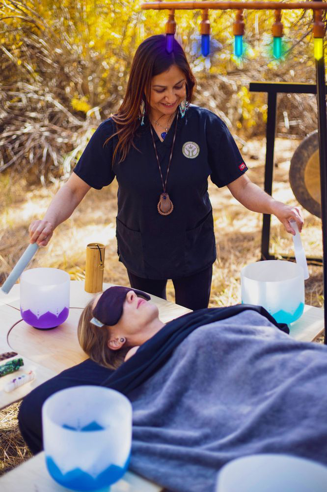 Practitioner performing a sound healing session outdoors with crystal singing bowls placed around the client on a massage table.