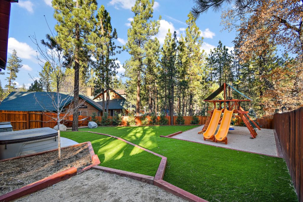 Backyard recreation area with playground, lawn, and hot tub surrounded by pine trees