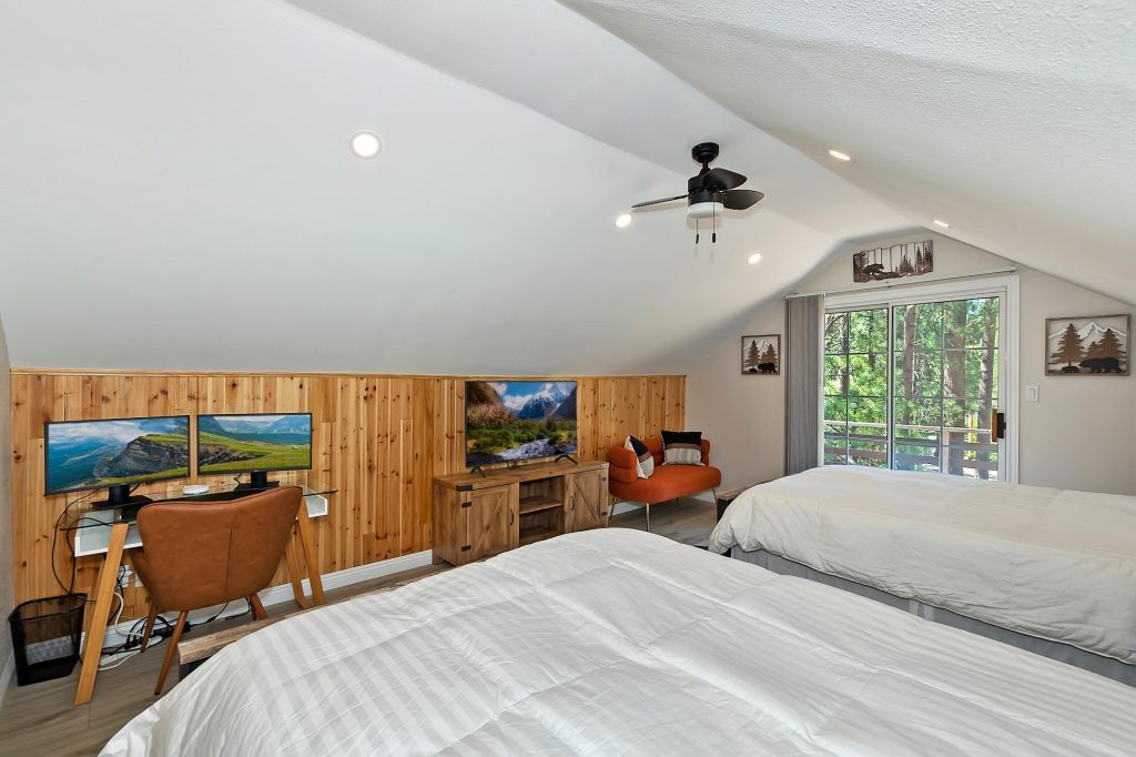 Bedroom with two beds, workspace with dual monitors, and a balcony overlooking pine trees
