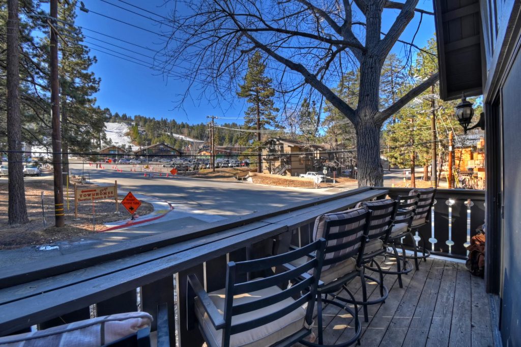 View from a balcony with chairs, overlooking a snowy ski slope, pine trees, and town buildings under a clear blue sky