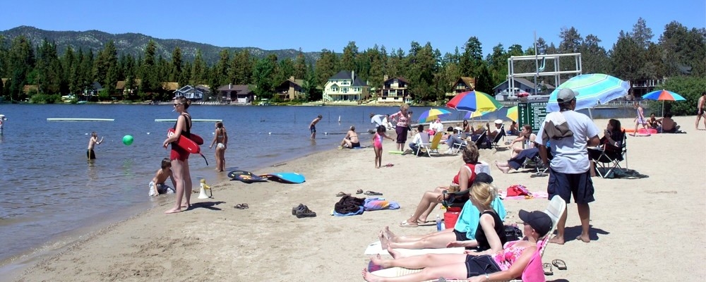 Busy lakeside beach area with visitors relaxing on the sand, swimming in the lake, umbrellas and beach gear set up along the shore with mountain and forest views in the background.
