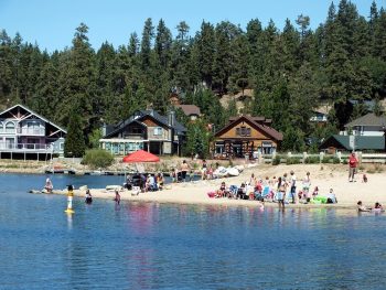 Families and visitors enjoying a sandy swimming beach on Big Bear Lake with calm water, forested mountains in the background, and cabins along the shoreline.