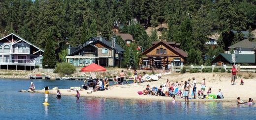 Families and visitors enjoying a sandy swimming beach on Big Bear Lake with calm water, forested mountains in the background, and cabins along the shoreline.
