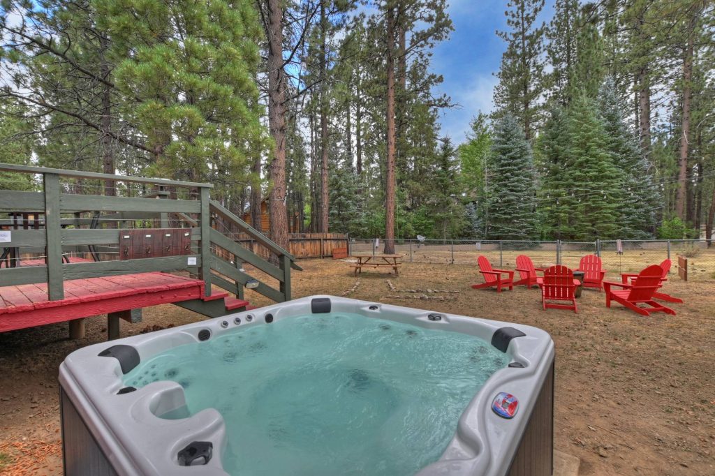 Outdoor hot tub beside the deck overlooking the fenced backyard with fire pit seating and tall pine trees