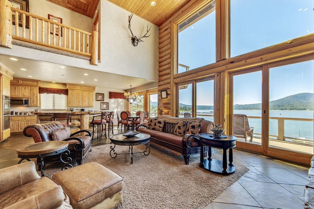 A living room with a brown couch and chairs, and a large wall of windows and sliding glass doors that open to a lake in Big Bear.