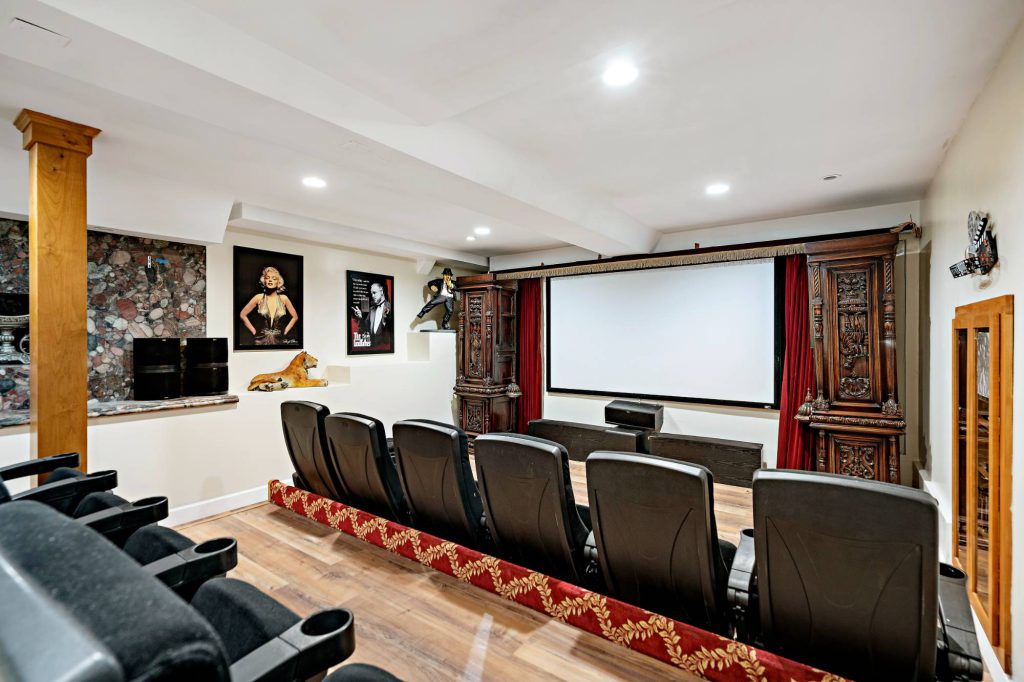 A cozy home theater in Big Bear with two rows of black leather seats facing a large screen, flanked by ornate wooden cabinets with red curtains.